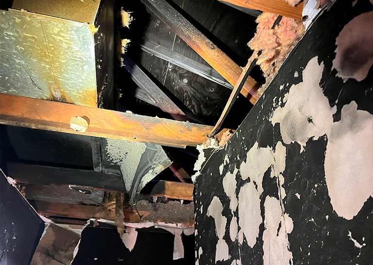 exposed-roof-beams-soot-cleanup Exposed roof beams and insulation with water damage, peeling paint, and decay, illuminated by a flashlight in a dark space.