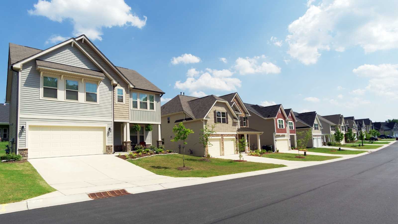 modern-suburban-houses A suburban street lined with modern houses featuring landscaped yards under a blue sky with fluffy clouds.