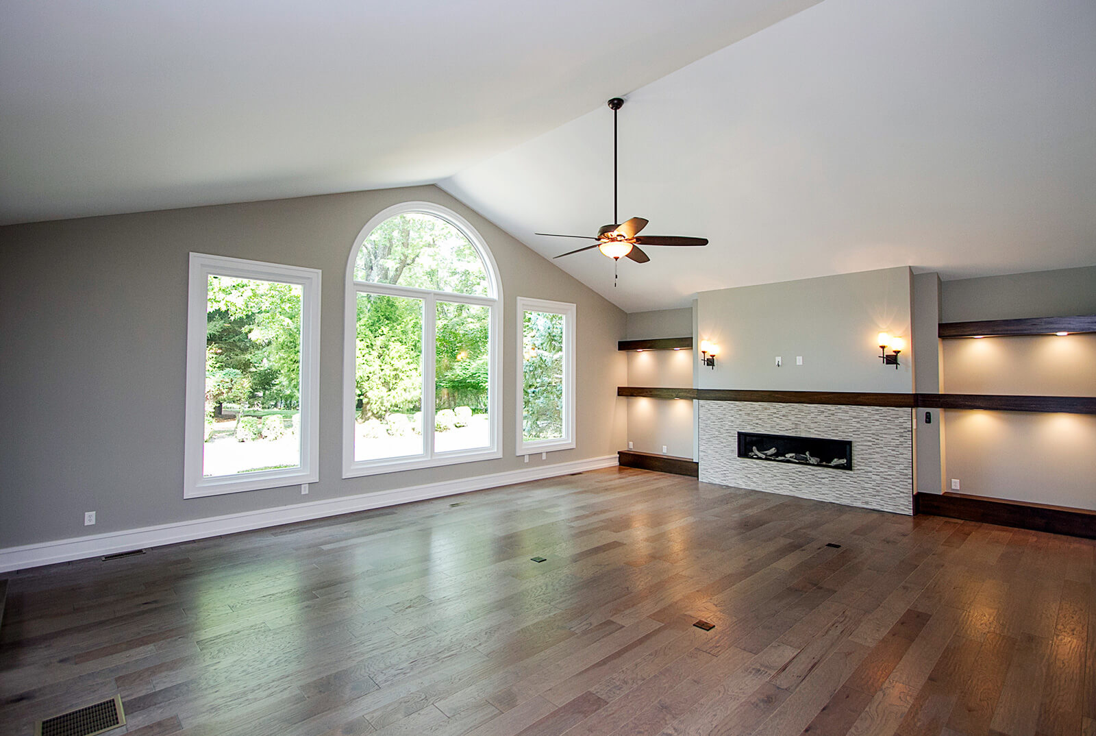 home-remodel-1 Modern living room with vaulted ceiling, large arched window, and rectangular windows overlooking green trees. The room features a light gray wall, wooden floor, a fireplace with a textured white tile surround, and dark wood mantels with recessed lighting.